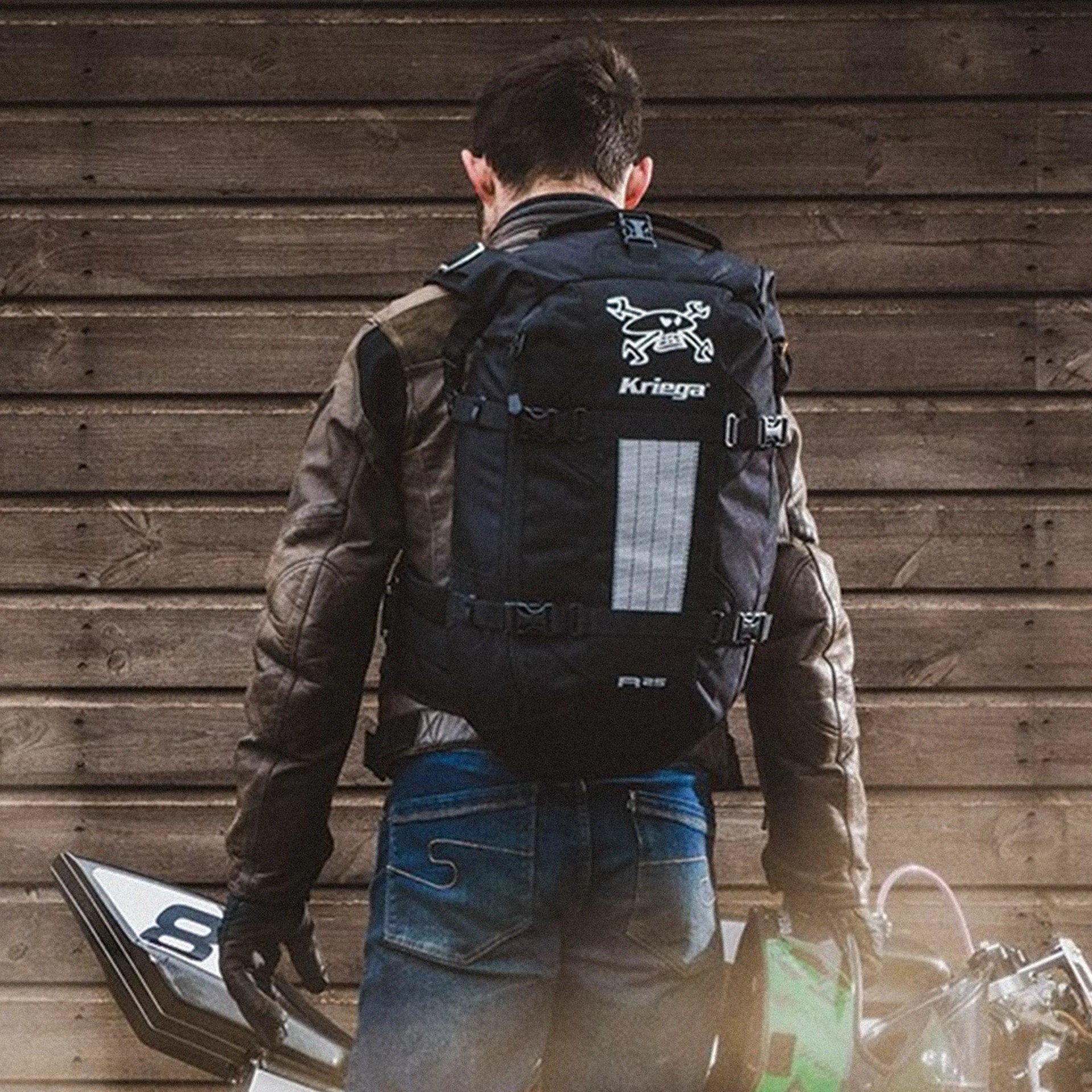 Person wearing a brown jacket and blue jeans with a black Kriega backpack, standing in front of a wooden wall.