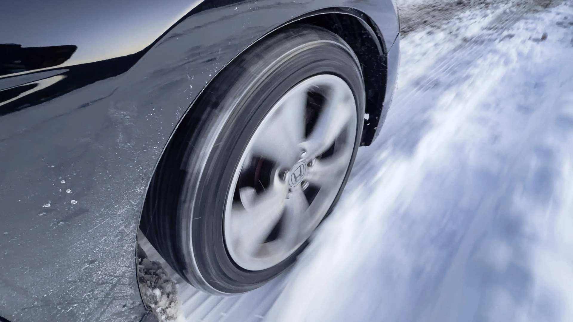 Spinning black car tire with silver alloy rim driving on snow-covered road.
