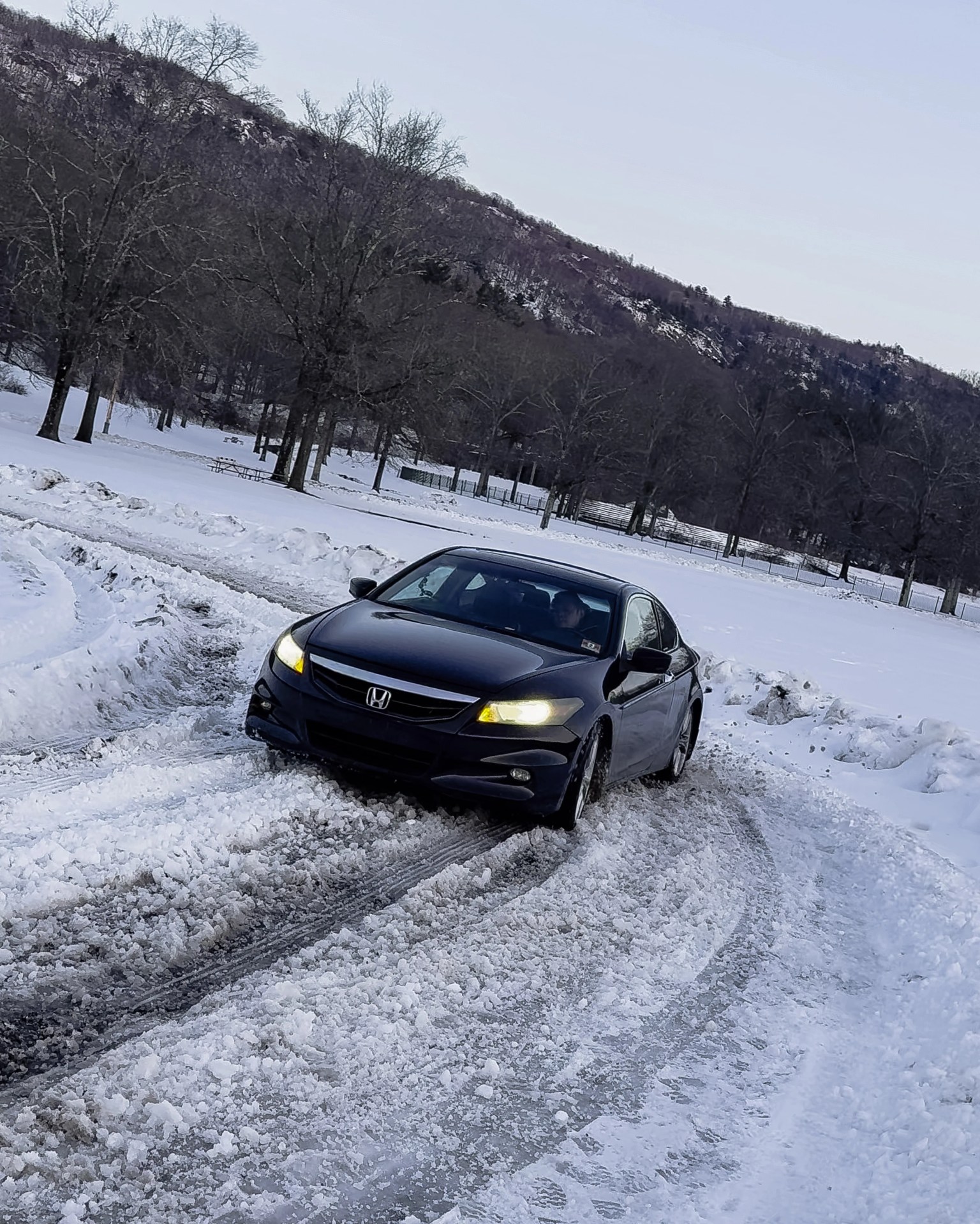 Black Honda sedan with headlights on driving on a snow-covered road near leafless trees and hills.
