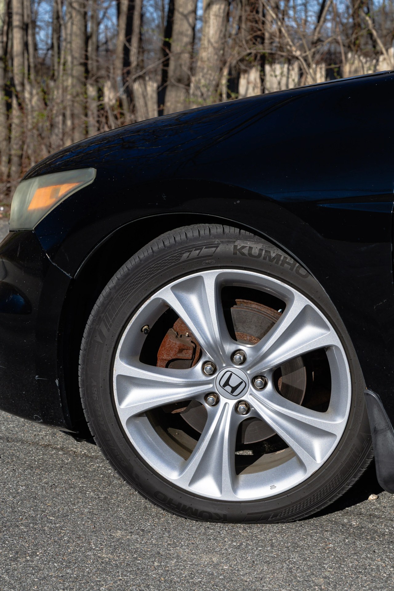 Close-up of a silver Honda alloy wheel with Kumho tire on a black car parked on asphalt.