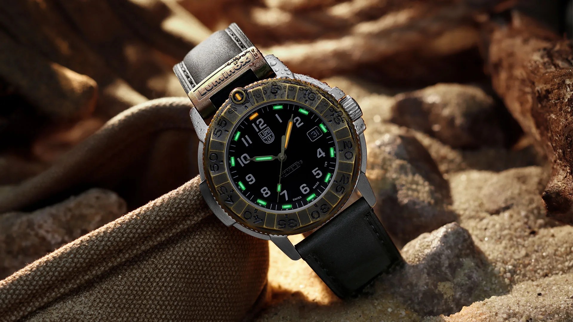 Black dial wristwatch with green and orange luminous markers, brass bezel, and black leather strap on sandy fabric.