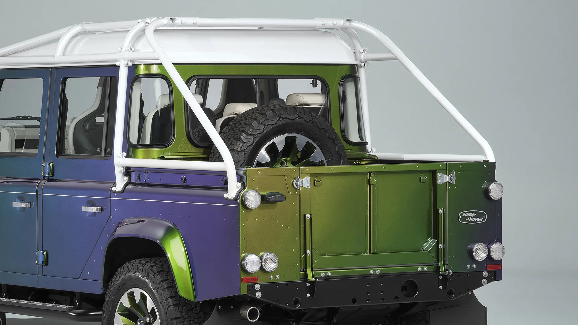 Rear side view of a two-tone blue and green Land Rover pickup with white roll cage and spare tire mounted inside the bed.