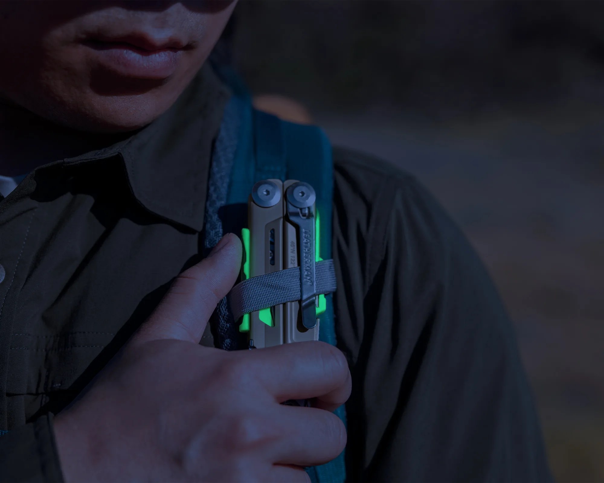 Close-up of a person holding a tan Leatherman multitool with green accents against a dark jacket.