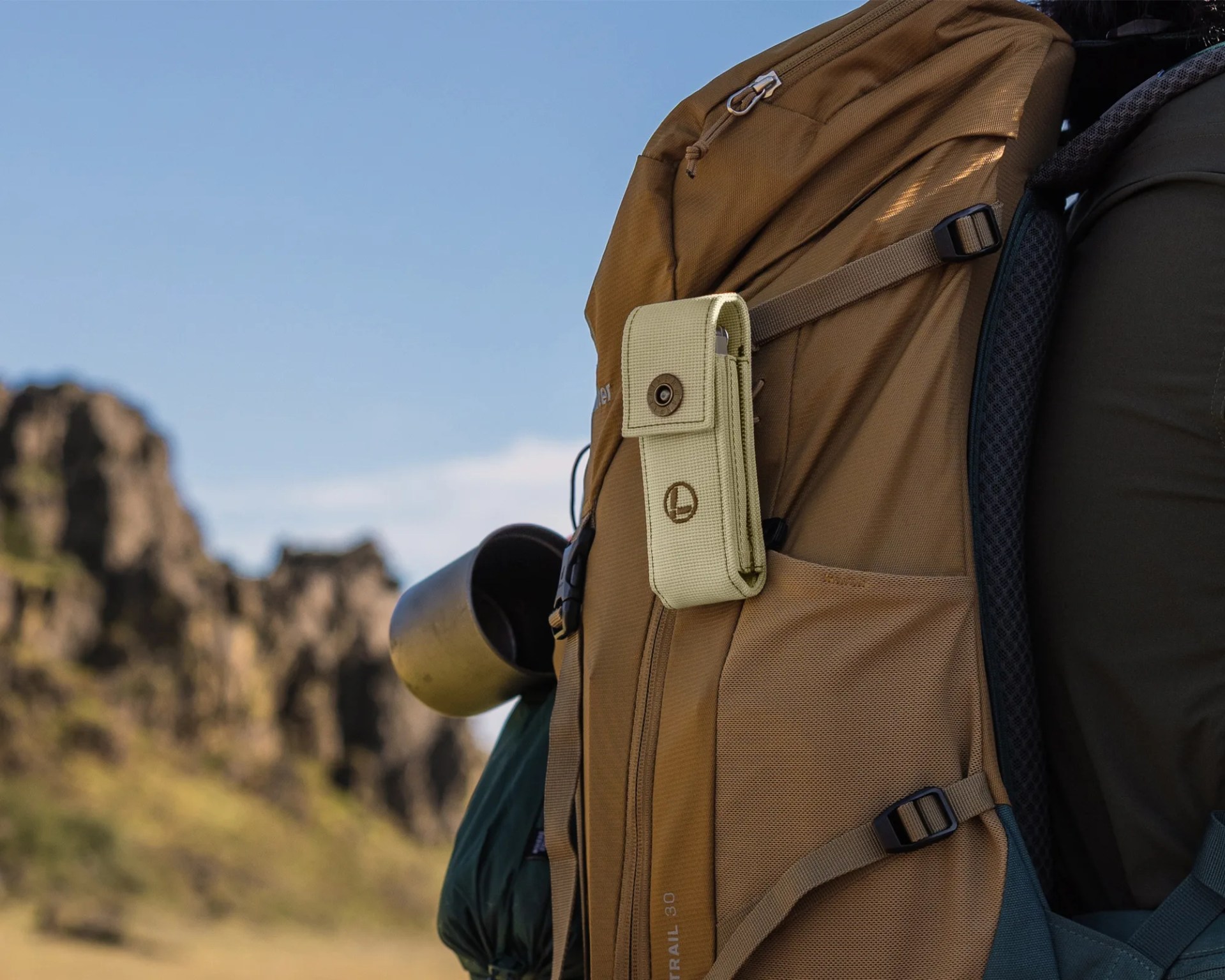 Tan and green hiking backpack with a beige strap pouch and a metal cup attached, set against a blurred outdoor background.