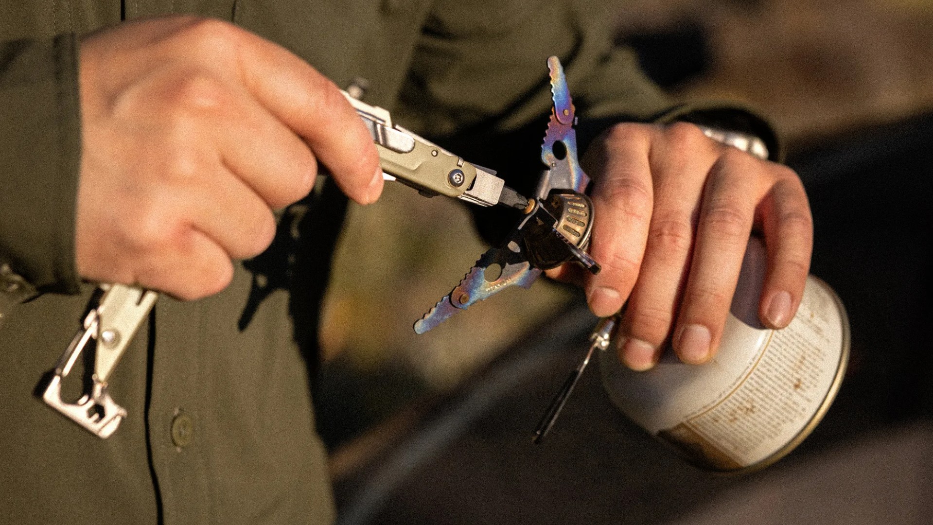 Hands holding a small camping stove with a multicolor metal burner and a silver multitool adjusting a part.