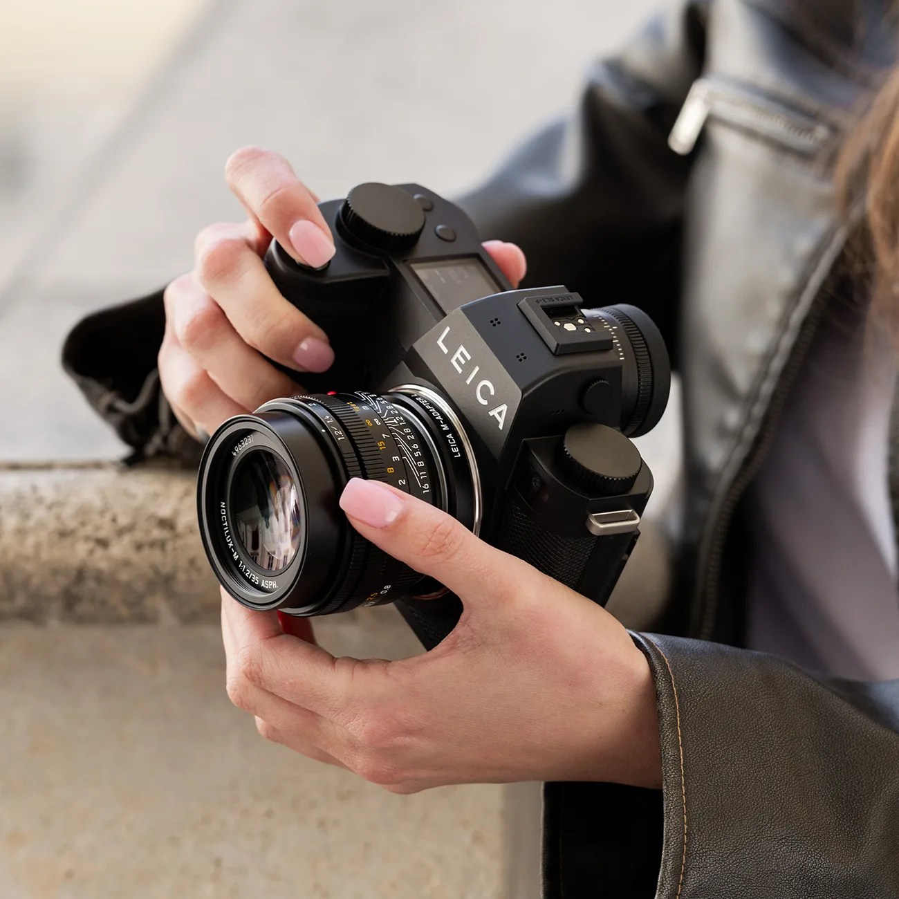 Black Leica camera with Noctilux-M 1:1.2/35 ASPH lens held in hands wearing a black leather jacket.