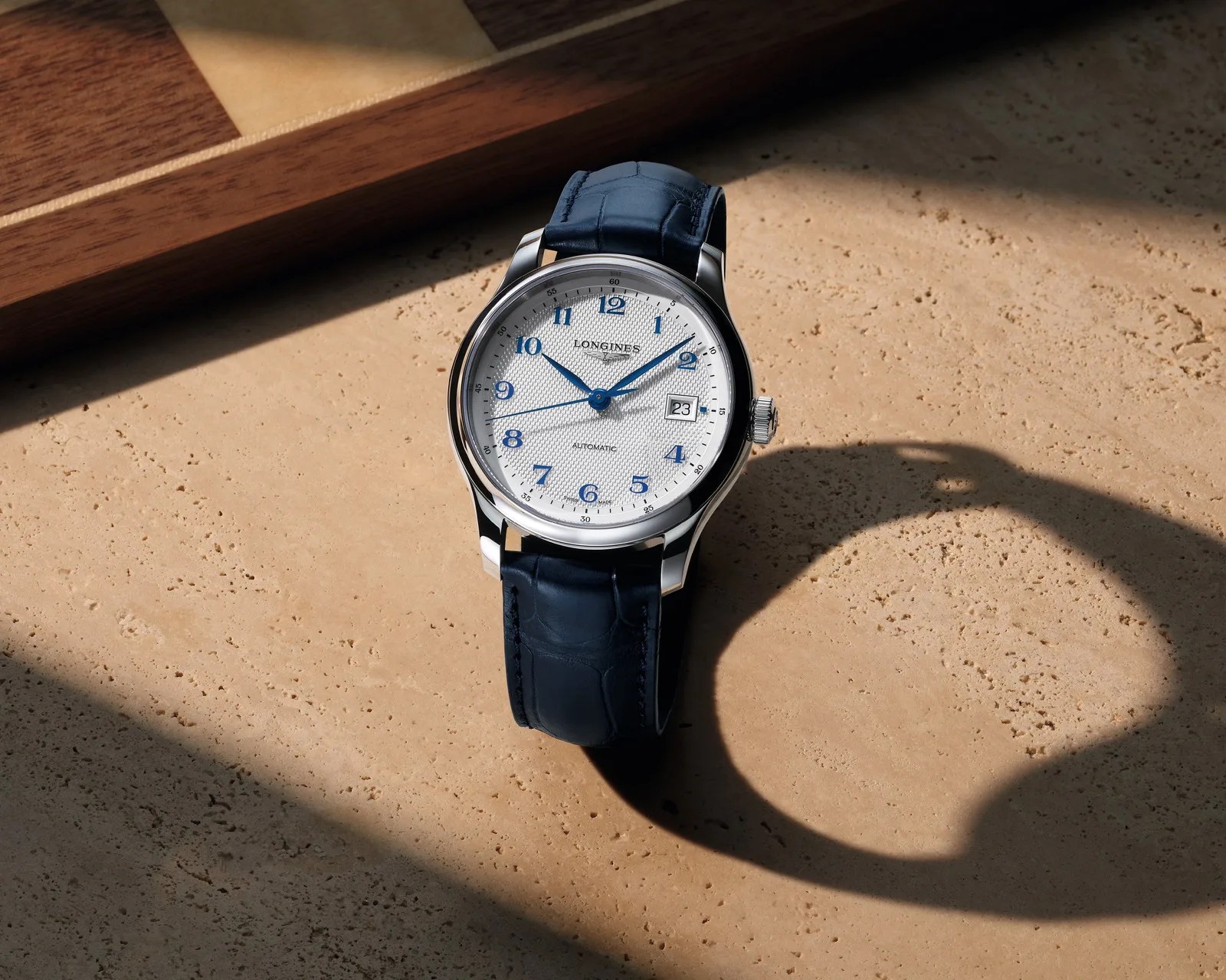 Silver Longines wristwatch with white textured dial, blue Arabic numerals, and dark blue leather strap on a stone surface.