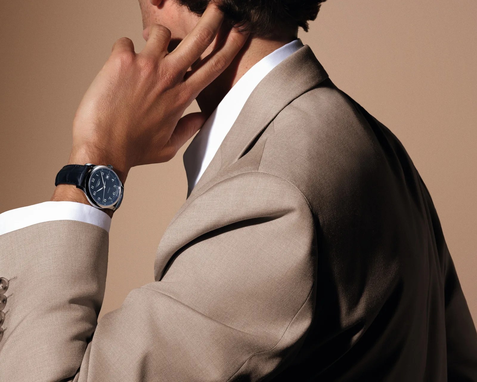 Man wearing a beige suit and white shirt with a blue leather strap wristwatch showing a blue dial.