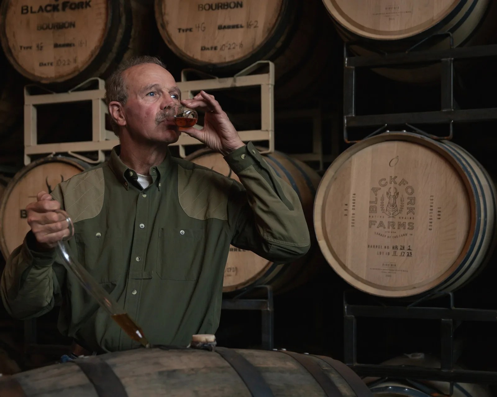 Man in an olive green shirt tasting bourbon in a glass in front of stacked wooden barrels labeled "Black Fork Farms.