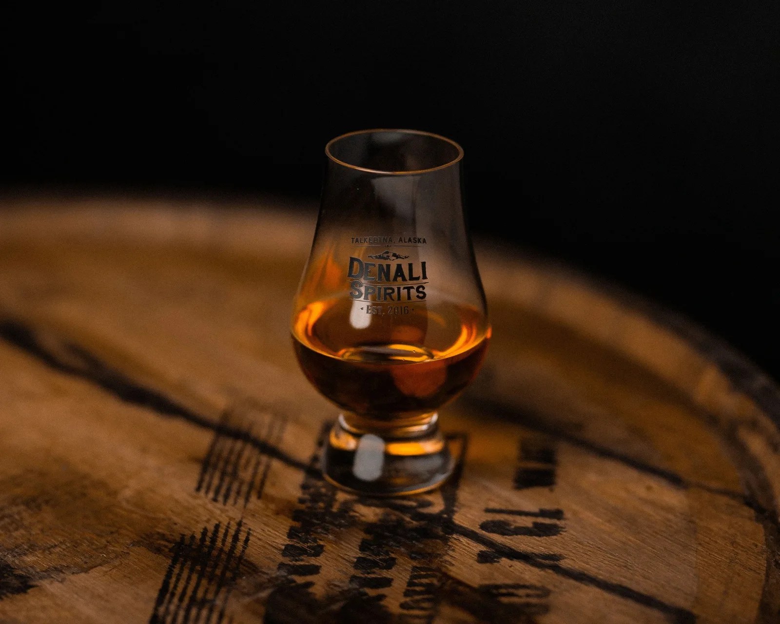 Whiskey glass with amber liquid labeled "Denali Spirits" on a wooden barrel top.