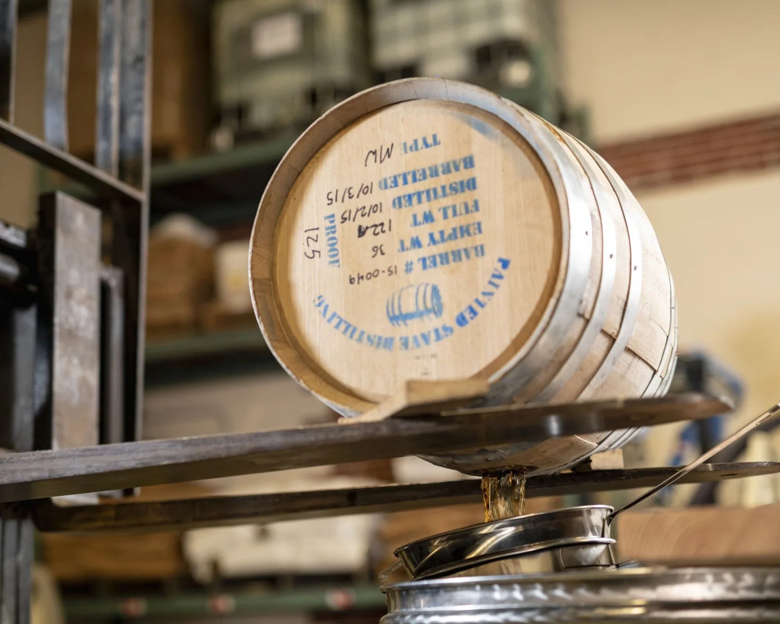Wooden barrel with blue text pouring liquid into a metal container in a warehouse.