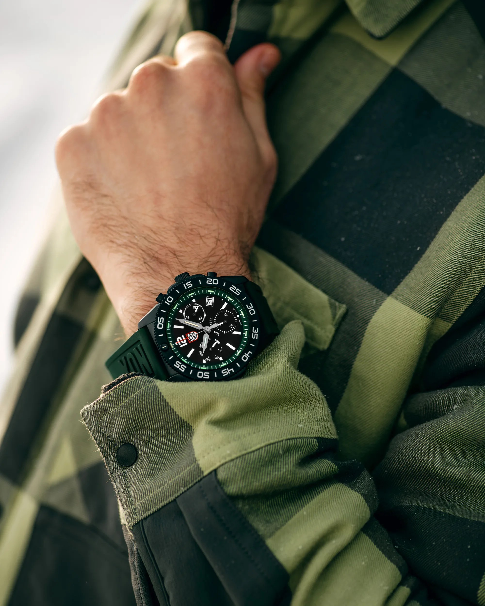 Black and green chronograph wristwatch with a black rubber strap worn on a wrist over a green and black checkered shirt.