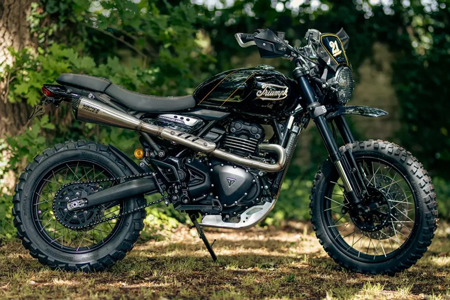 Black Triumph scrambler motorcycle with knobby tires and silver exhaust parked on grass near trees.
