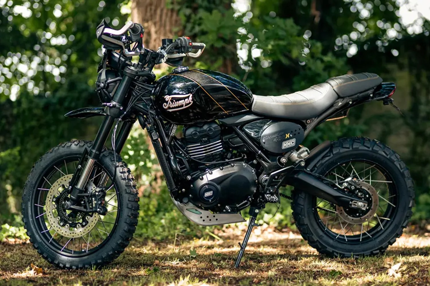 Black Triumph scrambler motorcycle with knobby tires parked on grass with trees in the background.