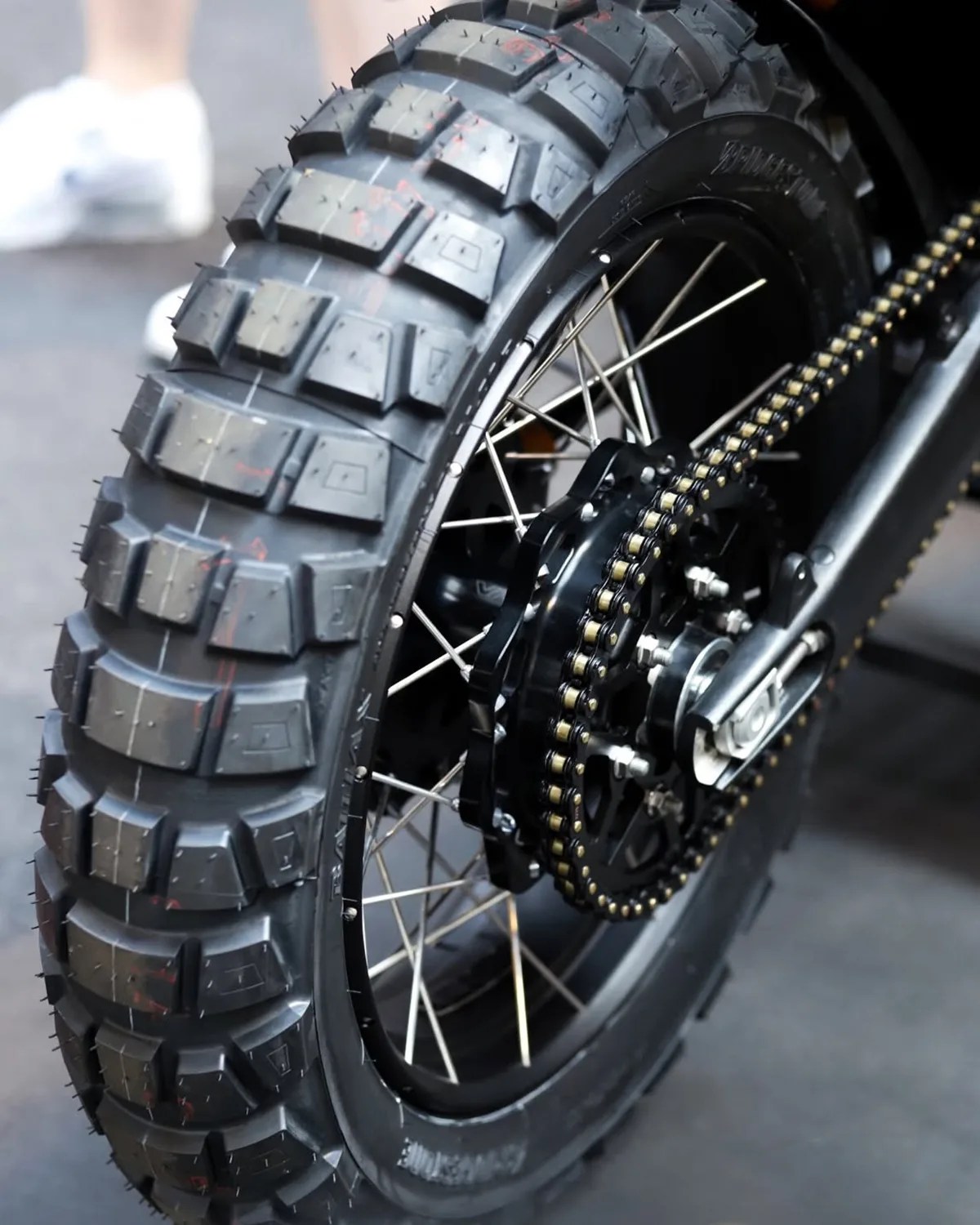 Close-up of a knobby off-road motorcycle tire with a black rim, silver spokes, and a gold-accented chain.