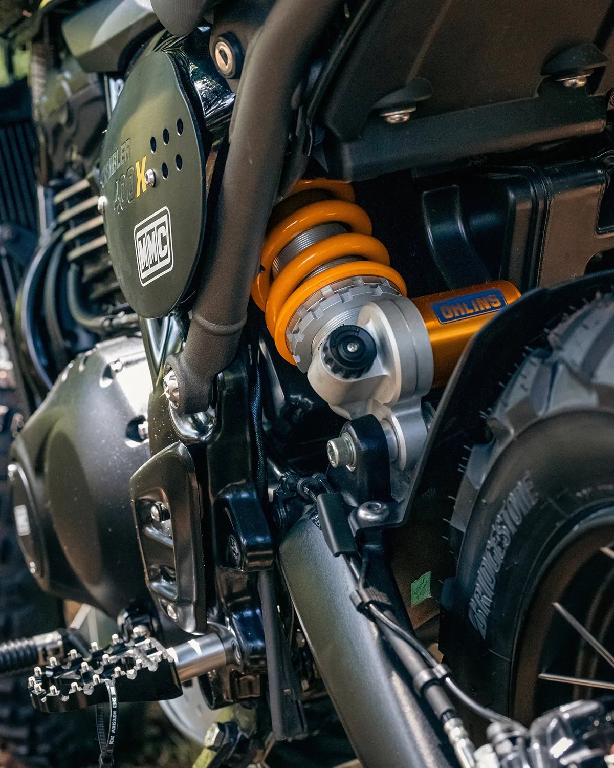 Close-up of a motorcycle's rear suspension with an orange &Ouml;hlins shock absorber and black MMC cover.