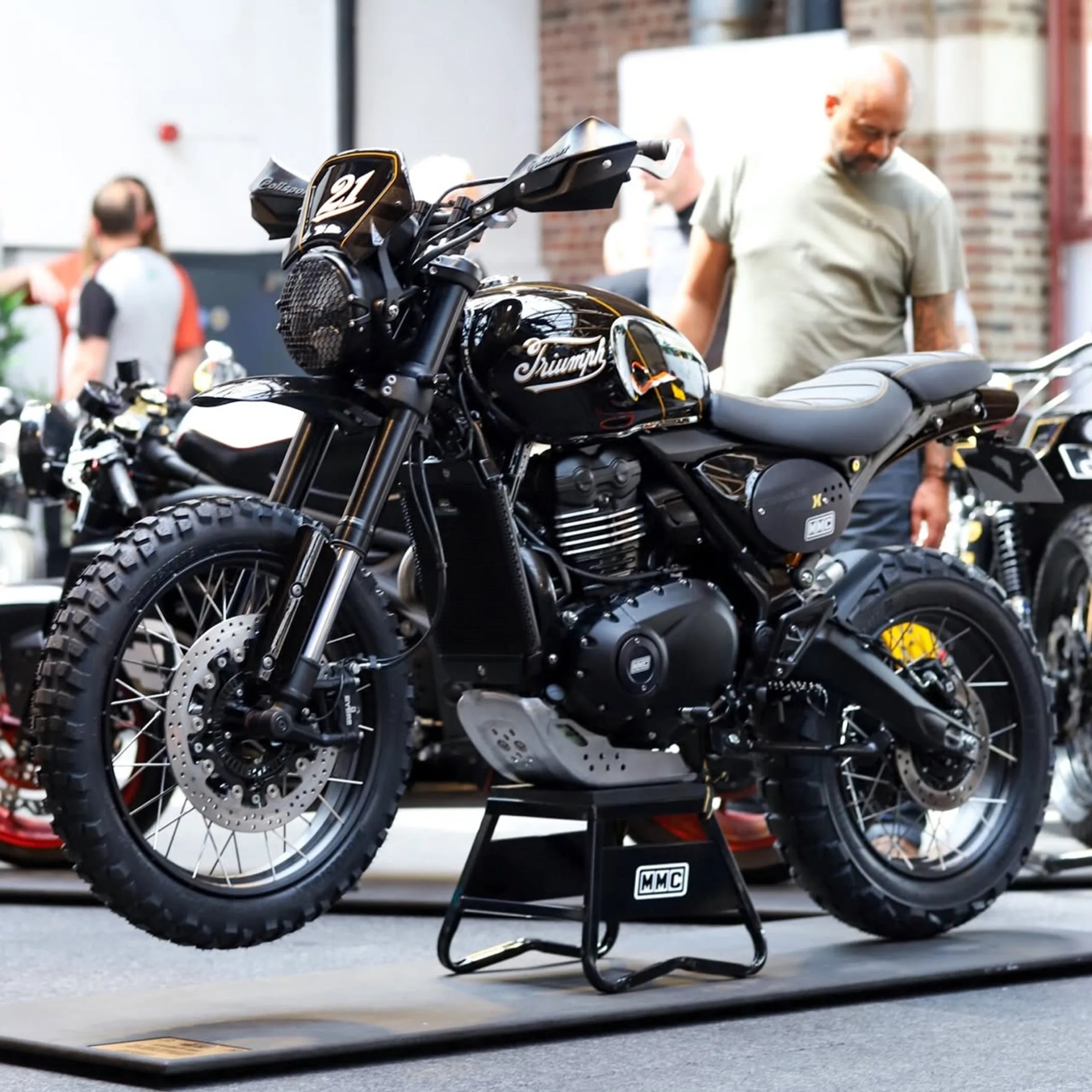 Black Triumph motorcycle with knobby tires displayed on a black MMC stand indoors.