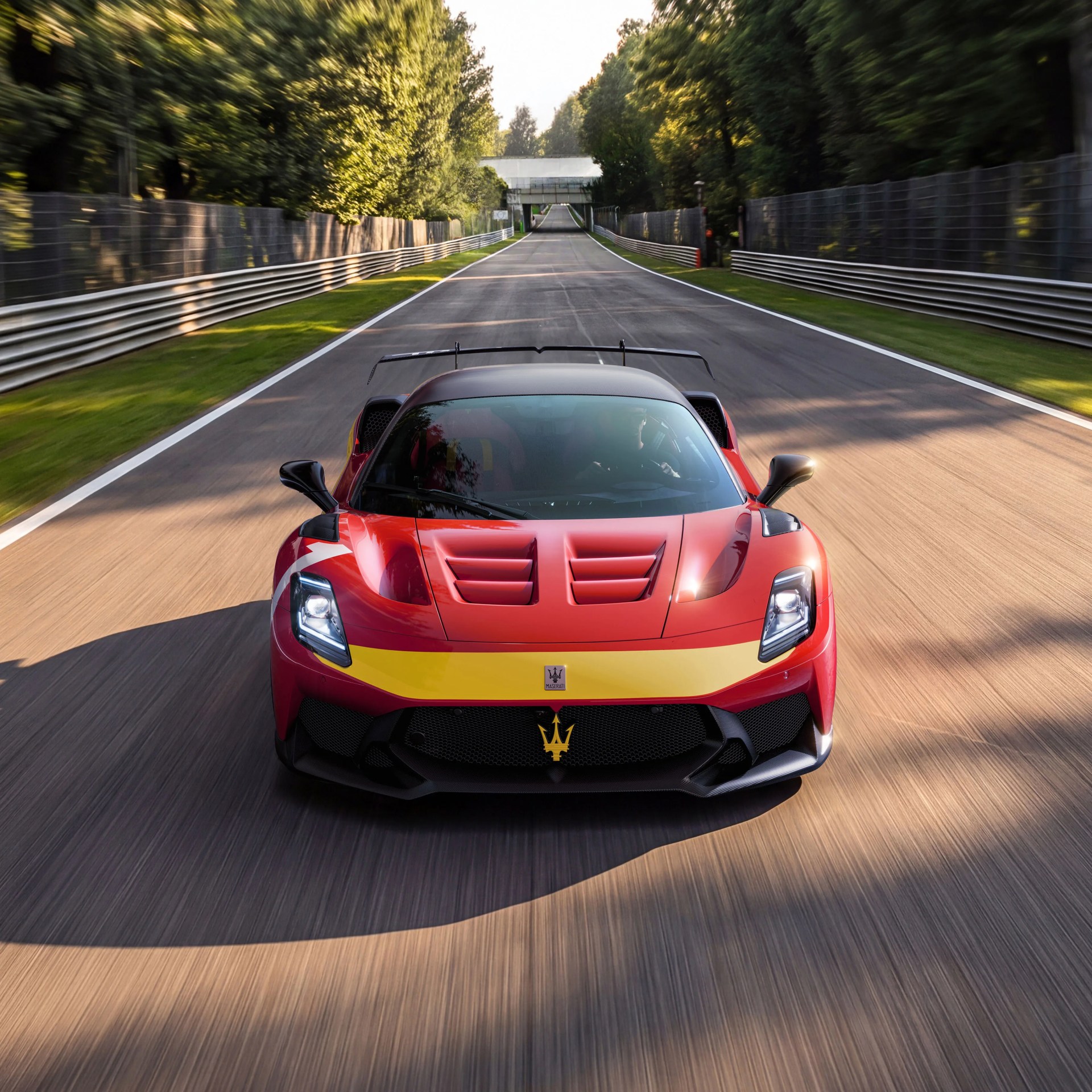 Red Maserati sports car with yellow stripe on a race track surrounded by trees.