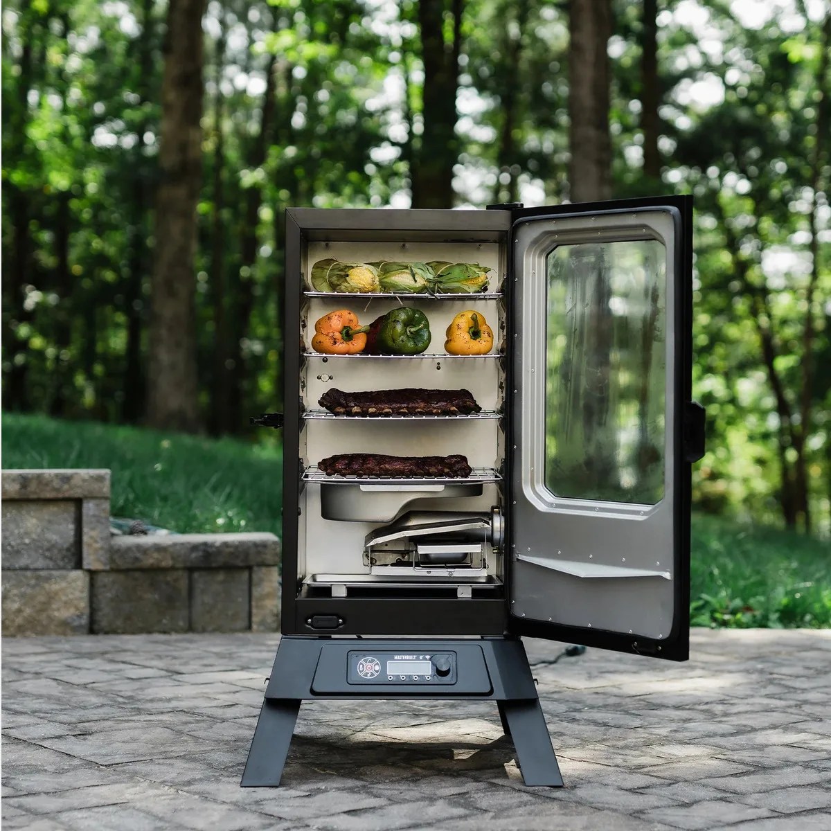 Black vertical electric smoker with open glass door showing corn, bell peppers, and ribs inside on a stone patio with trees in the background.