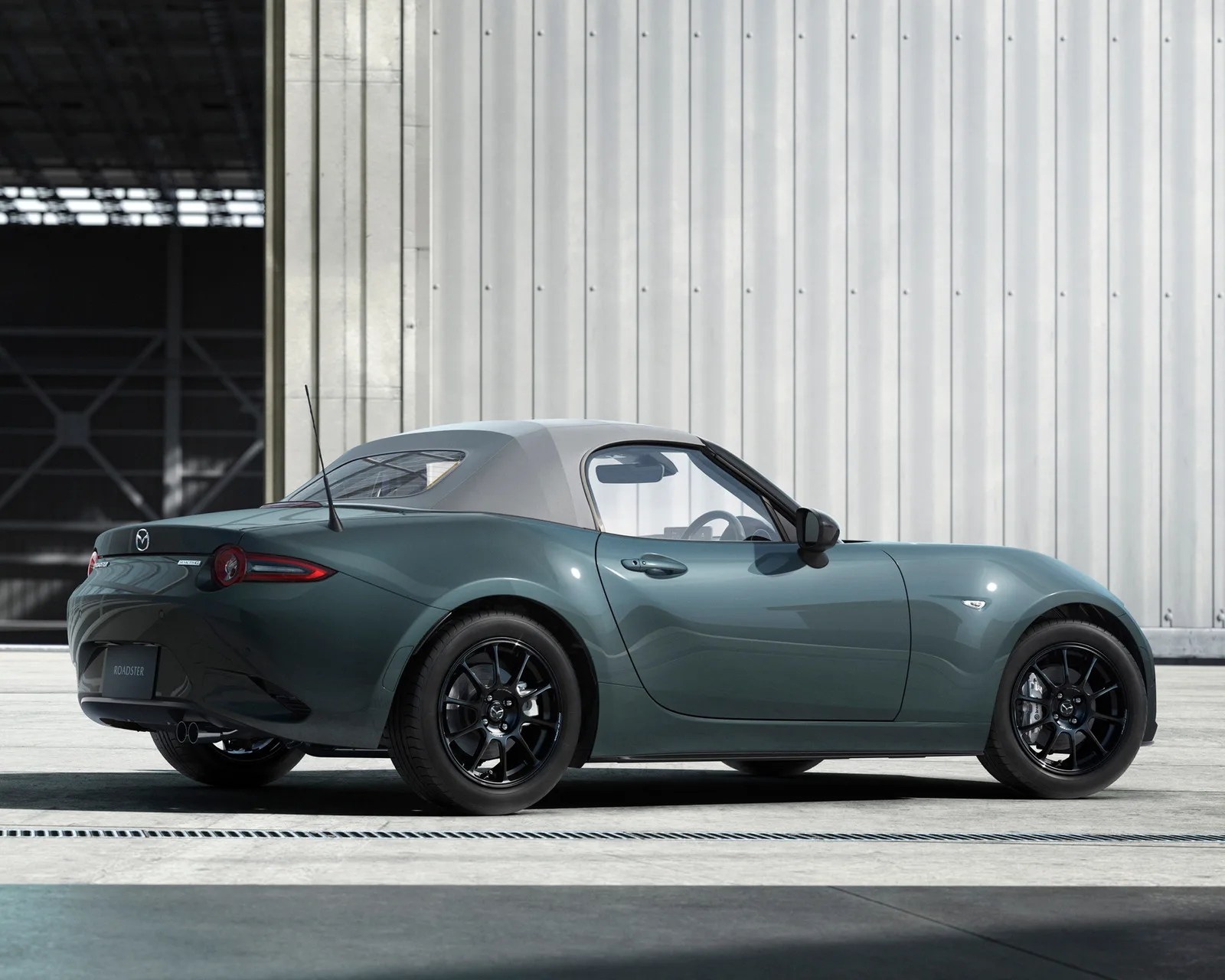 Green Mazda MX-5 Miata roadster with black wheels and gray soft top parked in front of a corrugated metal wall.