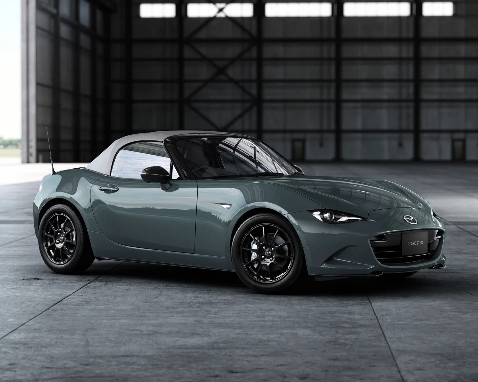 Glossy dark green Mazda MX-5 Miata roadster with black wheels and white soft top in a large industrial garage.