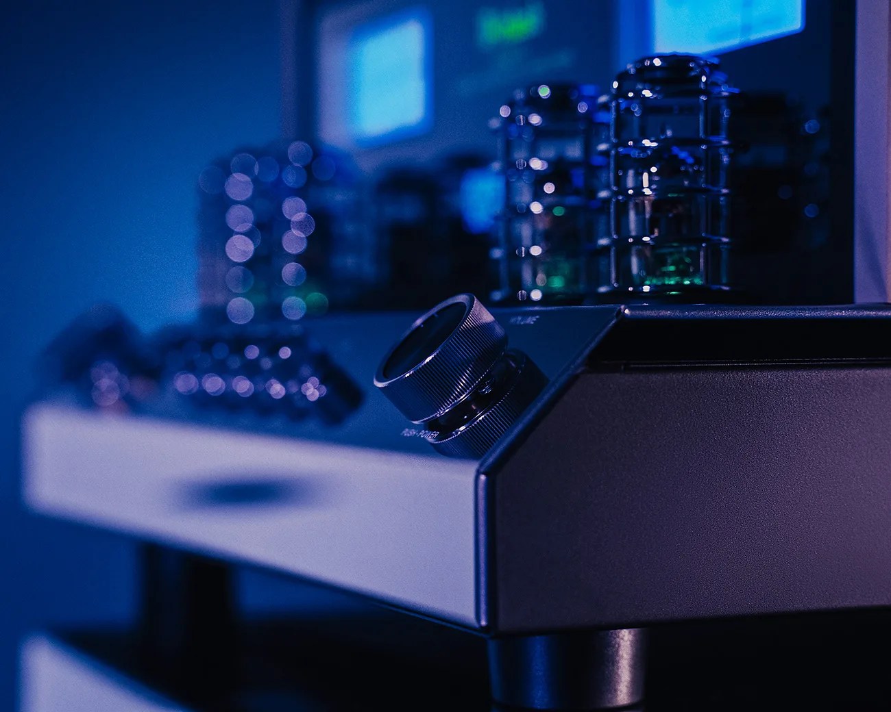 Close-up of a black audio amplifier knob with vacuum tubes glowing in blue light in the background.