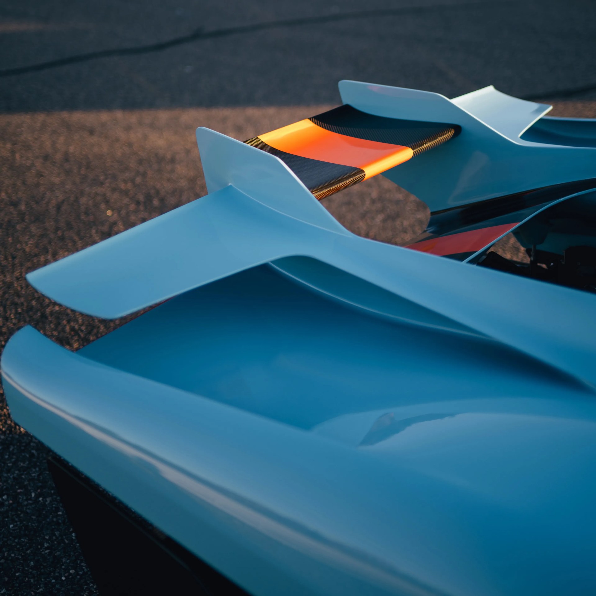 Glossy light blue car rear wing with carbon fiber and orange accent on asphalt surface.