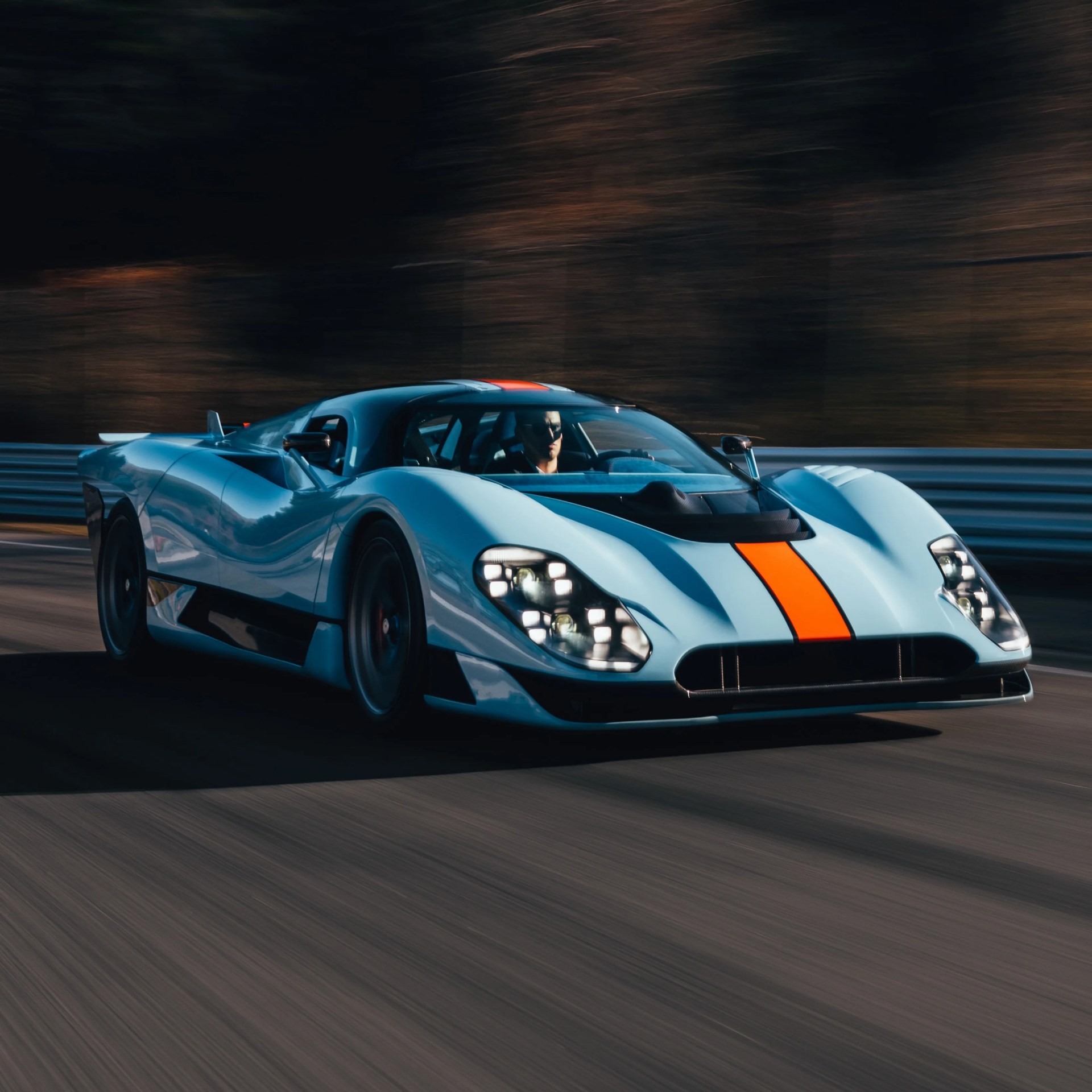 Light blue sports car with an orange racing stripe driving on a road with blurred background.