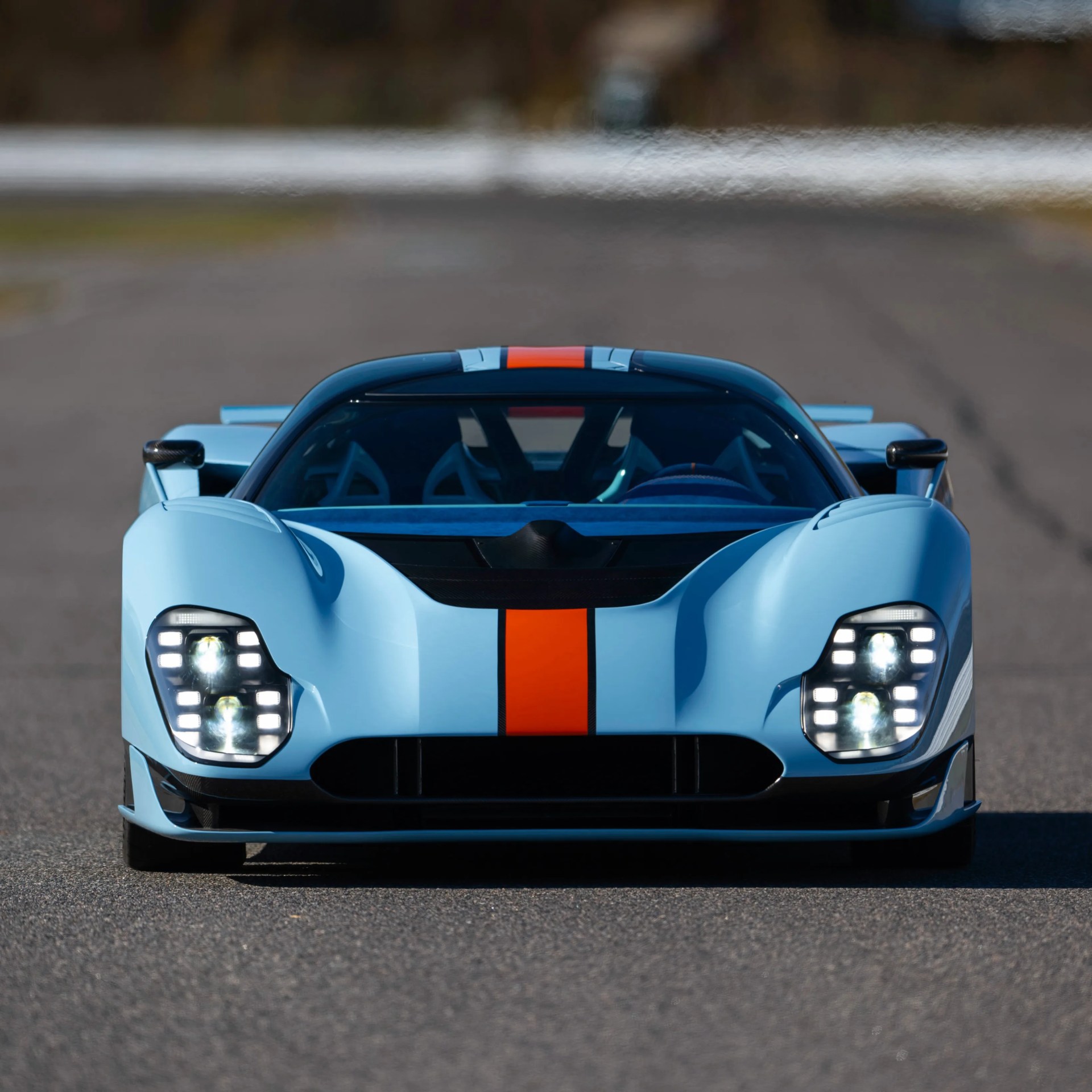 Light blue sports car with a central orange and black racing stripe, shown from the front on a road.