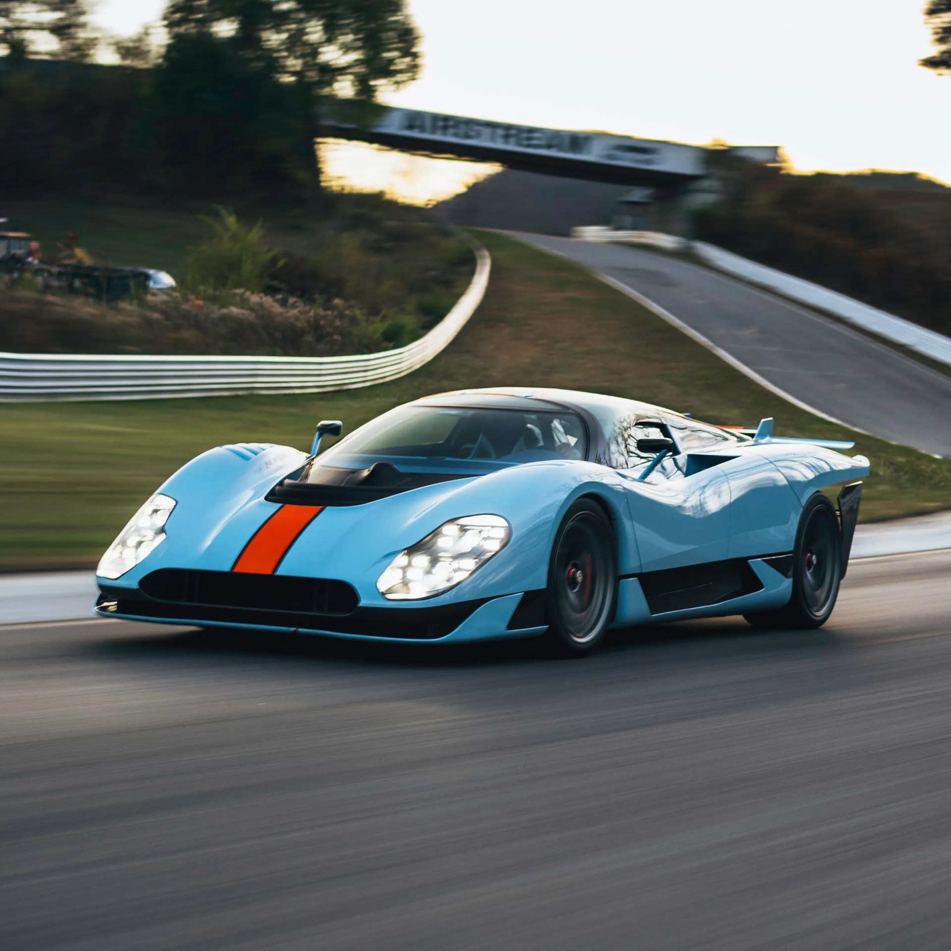 Light blue race car with an orange stripe driving on a racetrack near a bridge with "AIRSTREAM" signage.