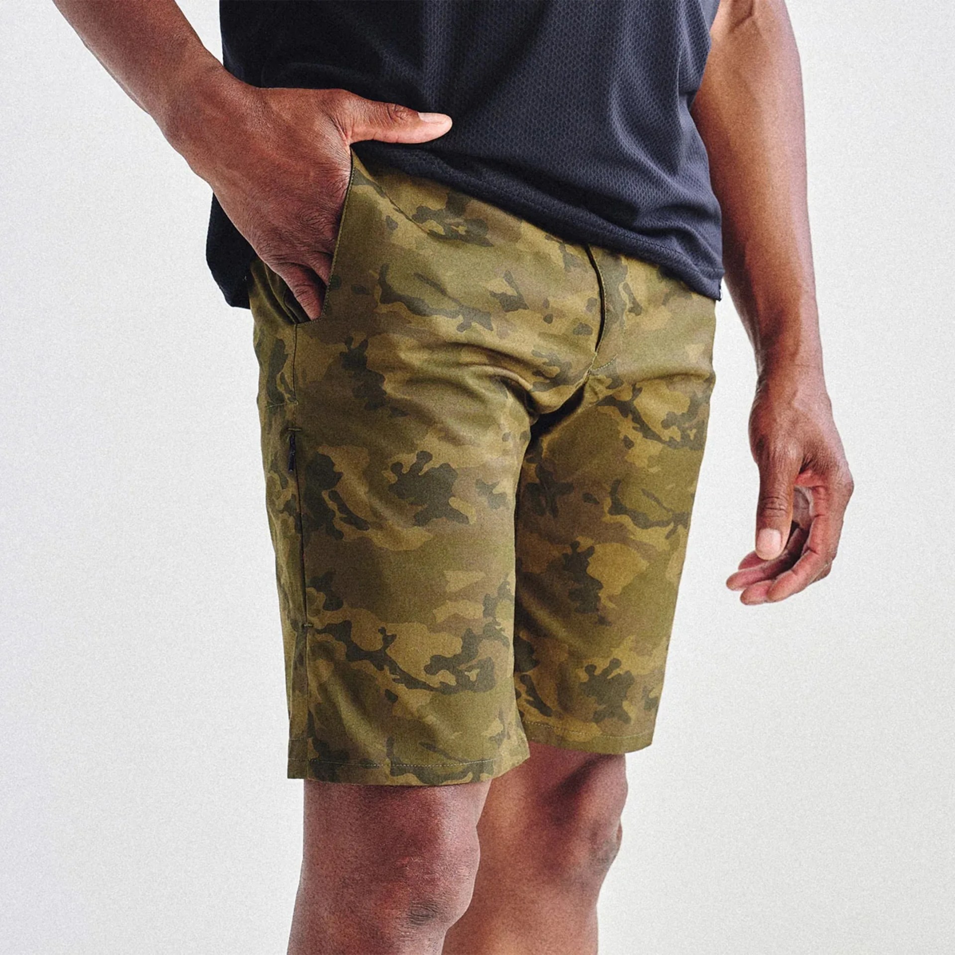 Man wearing olive green camouflage shorts and a black textured shirt with one hand in the pocket.