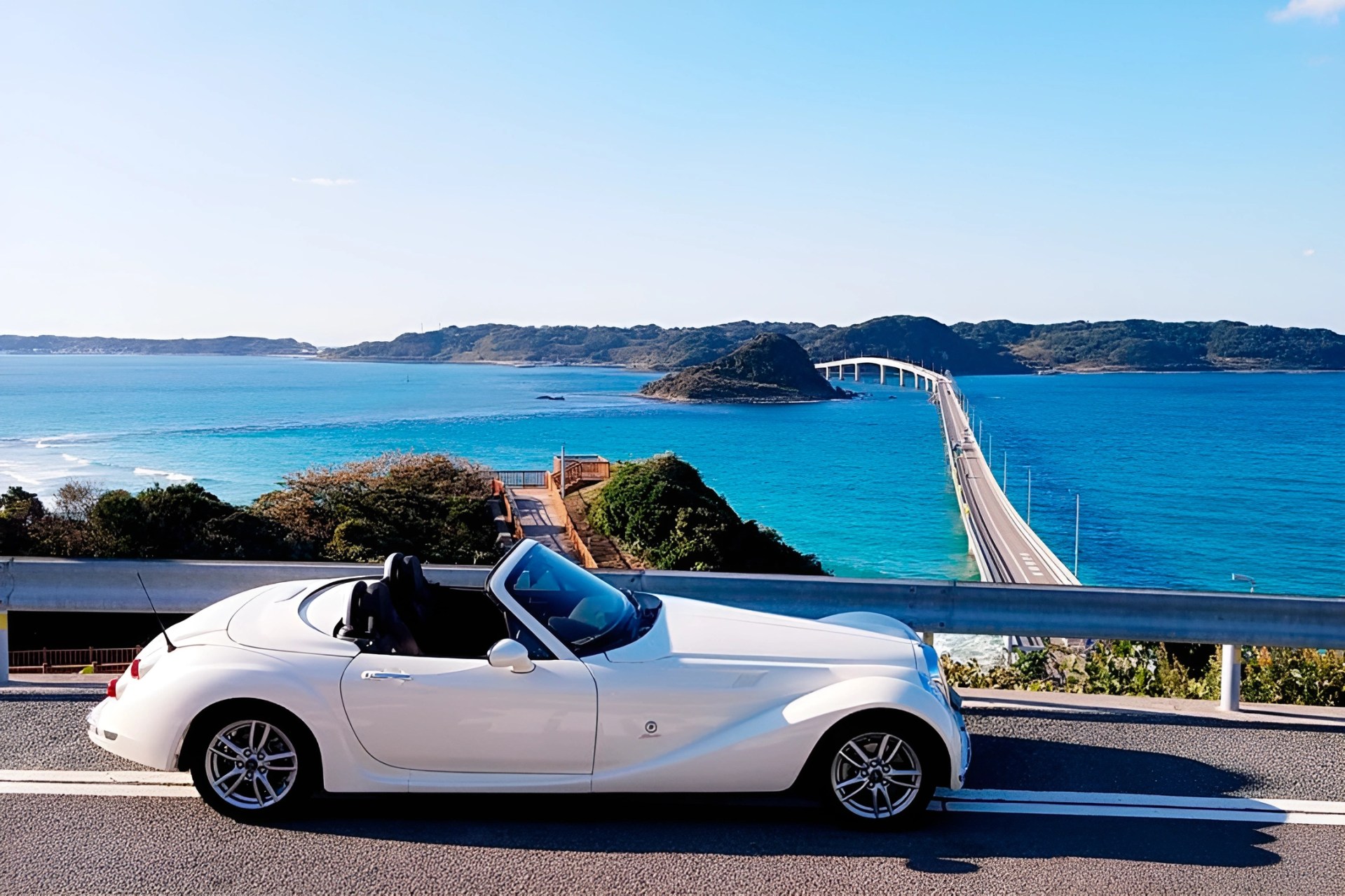White convertible sports car parked on a coastal road with a long bridge and blue ocean in the background.