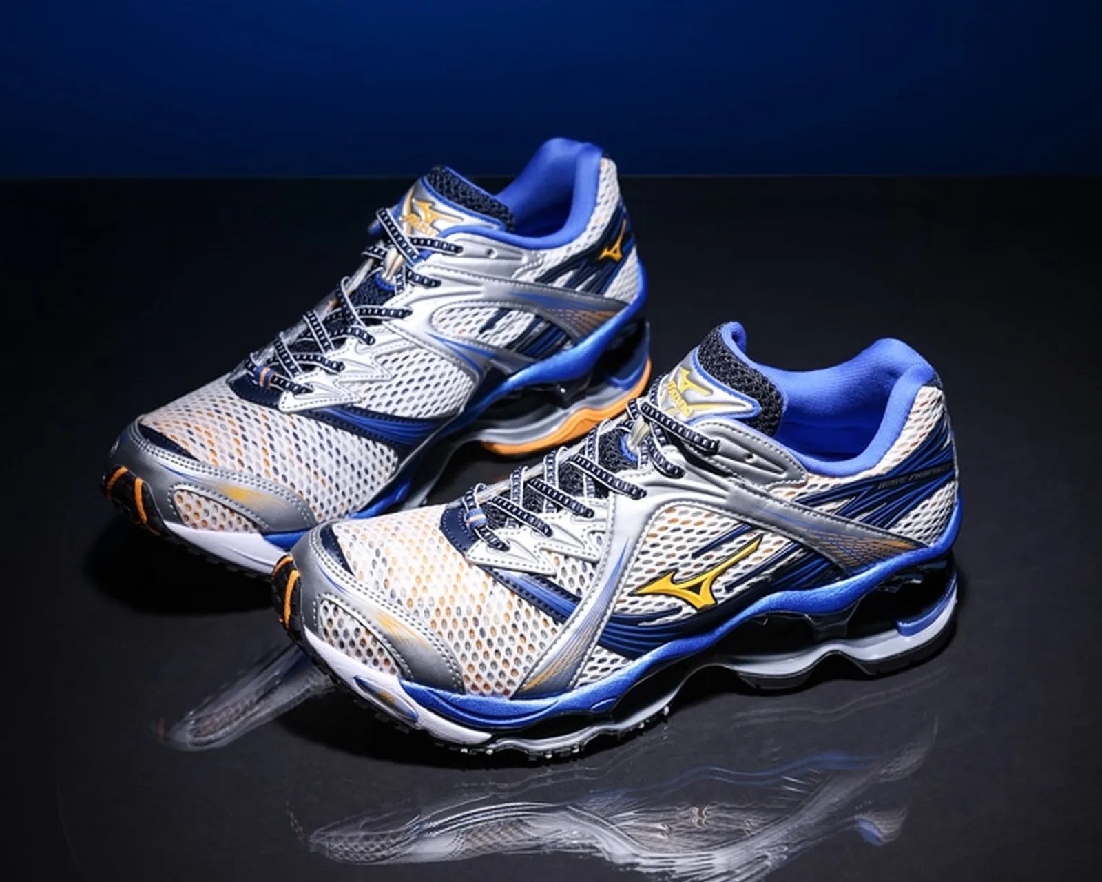 Pair of white, blue, and silver Mizuno running shoes with yellow logo on a reflective black surface.