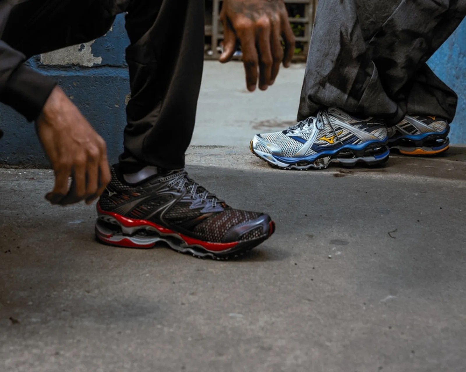 Two pairs of Mizuno running shoes, one black with red accents and the other white with blue and yellow accents, worn by people standing on concrete.