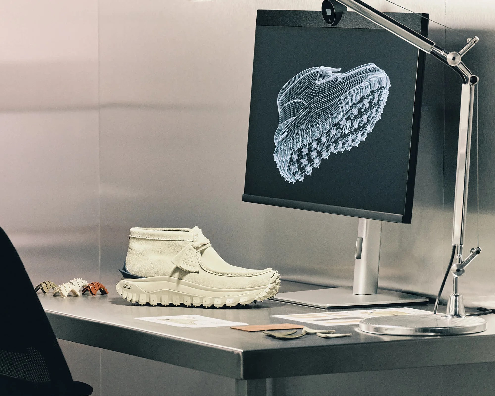 Beige high-top shoe on a desk next to a monitor displaying a wireframe 3D model of the shoe sole.