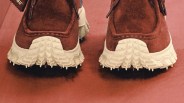 Brown suede shoes with thick, textured white soles shown from the back on a brown surface.