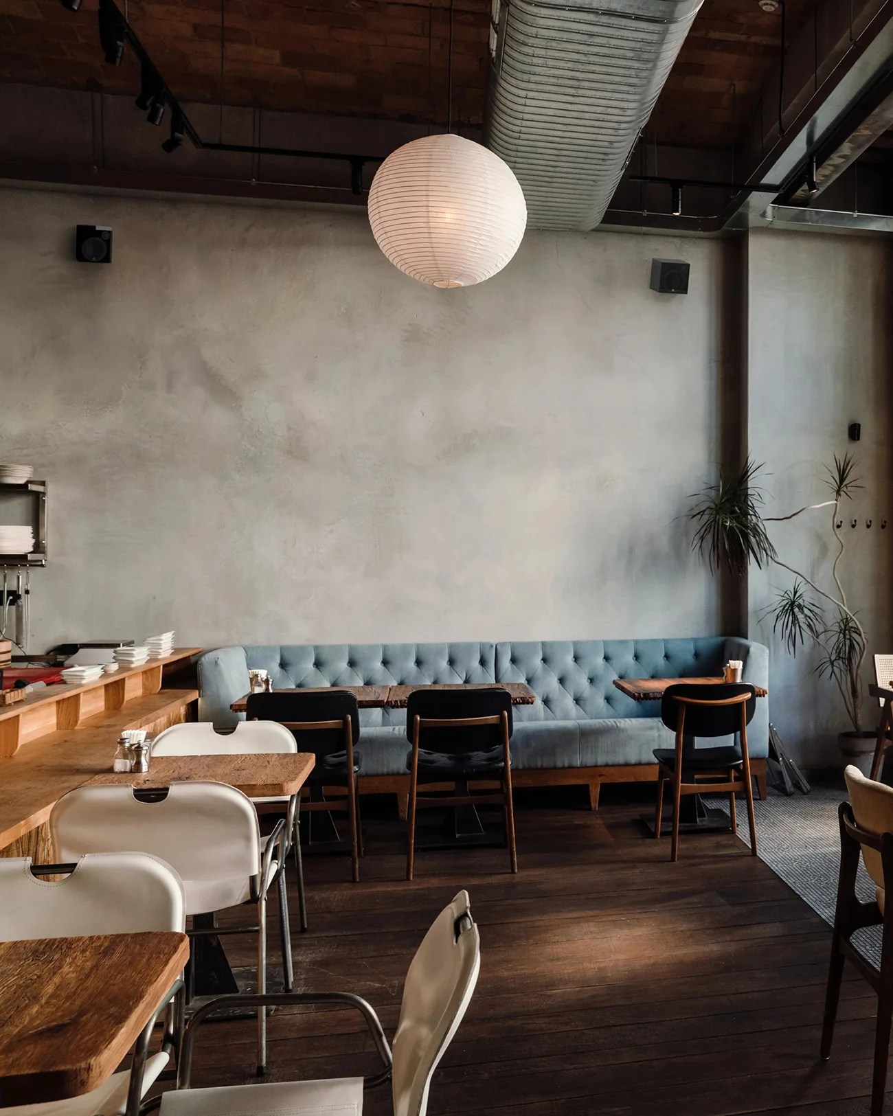 Industrial-style cafe interior with wooden tables, white and black chairs, a blue tufted bench, and a round paper lantern.