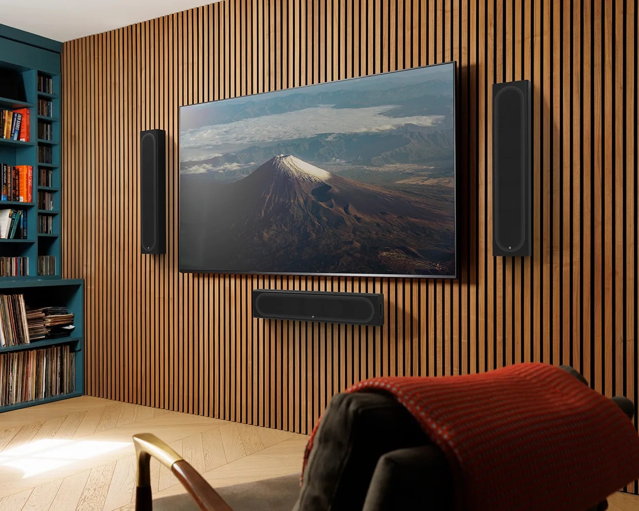 Wall-mounted flat-screen TV displaying a mountain scene with three black speakers on a wooden slat wall.