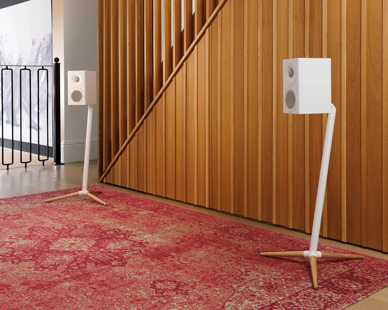 Pair of white bookshelf speakers on white and wood tripod stands placed on a red patterned rug in front of a wooden slatted wall.