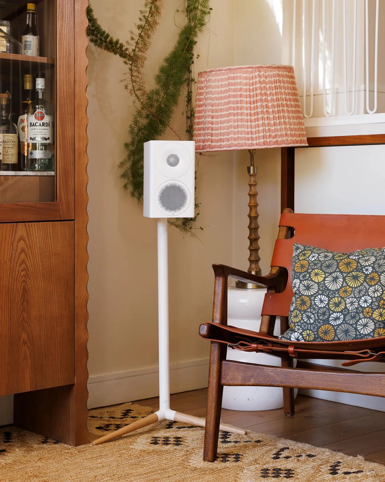 White speaker on a white and wood tripod stand next to a wooden chair with a patterned cushion and a lamp with a pink shade.