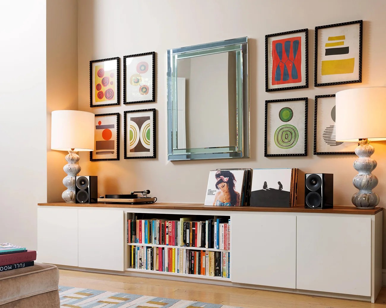 White low cabinet with wooden top holding two marble lamps, black speakers, a turntable, and vinyl records, below framed abstract art and a rectangular mirror.