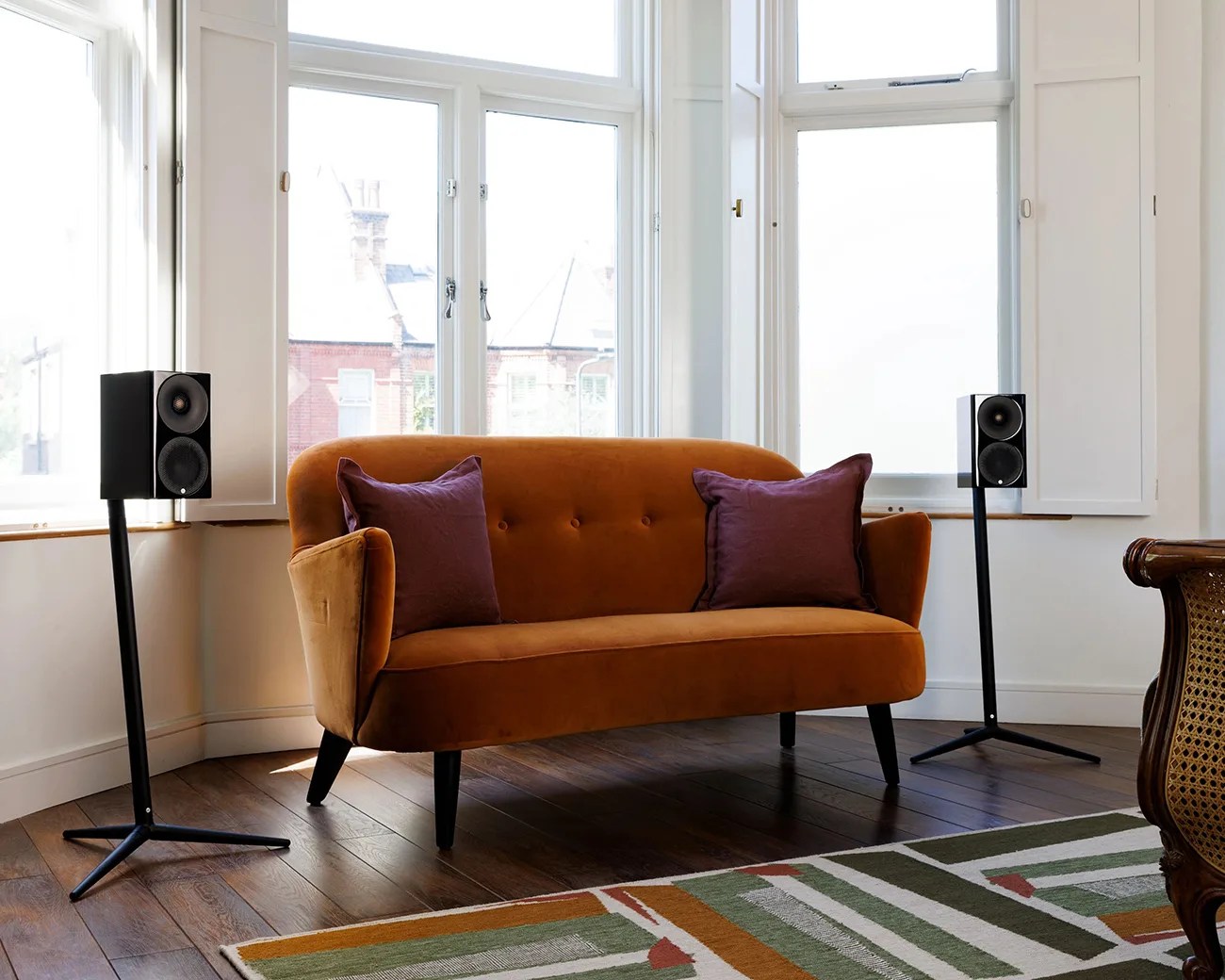 Rust-colored velvet loveseat with two purple pillows between two black speakers on stands in a bright room.
