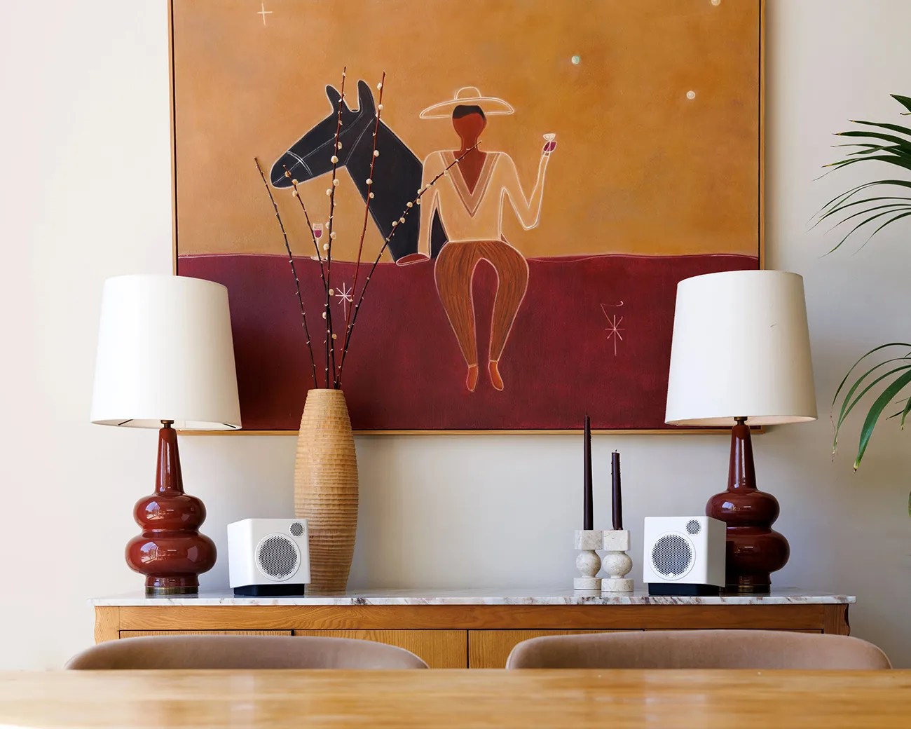 Wooden sideboard with marble top holding two brown ceramic lamps with white shades, a tall ribbed vase with branches, two white cube speakers, and two black candles in stone holders, below a large abstract painting of a person with a horse.
