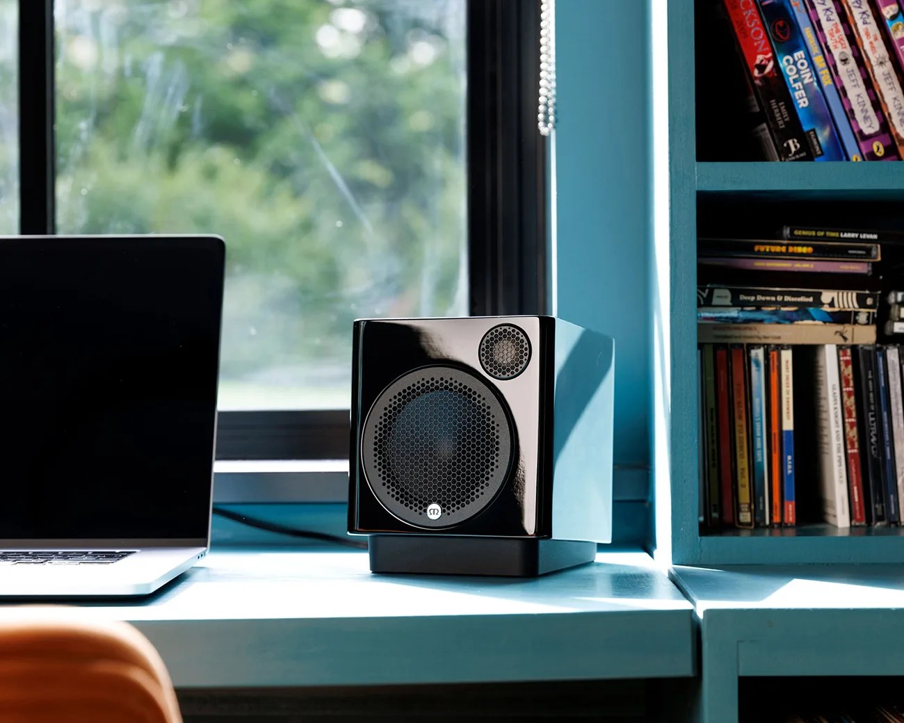 Glossy black cube speaker with metal mesh grille on a blue desk near a window and a laptop.