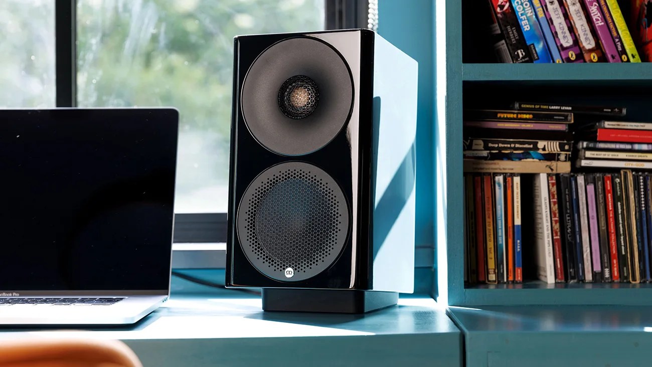 Glossy black and white bookshelf speaker on a blue desk next to a closed laptop and a bookshelf.