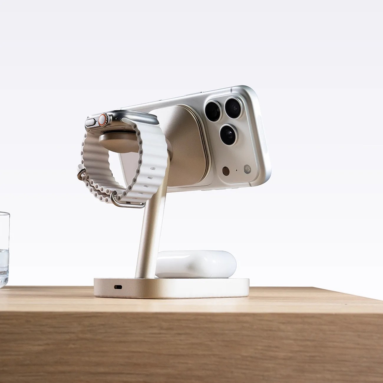 Silver multi-device charging stand holding a silver smartphone, a smartwatch with a white band, and white wireless earbuds case on a wooden surface.
