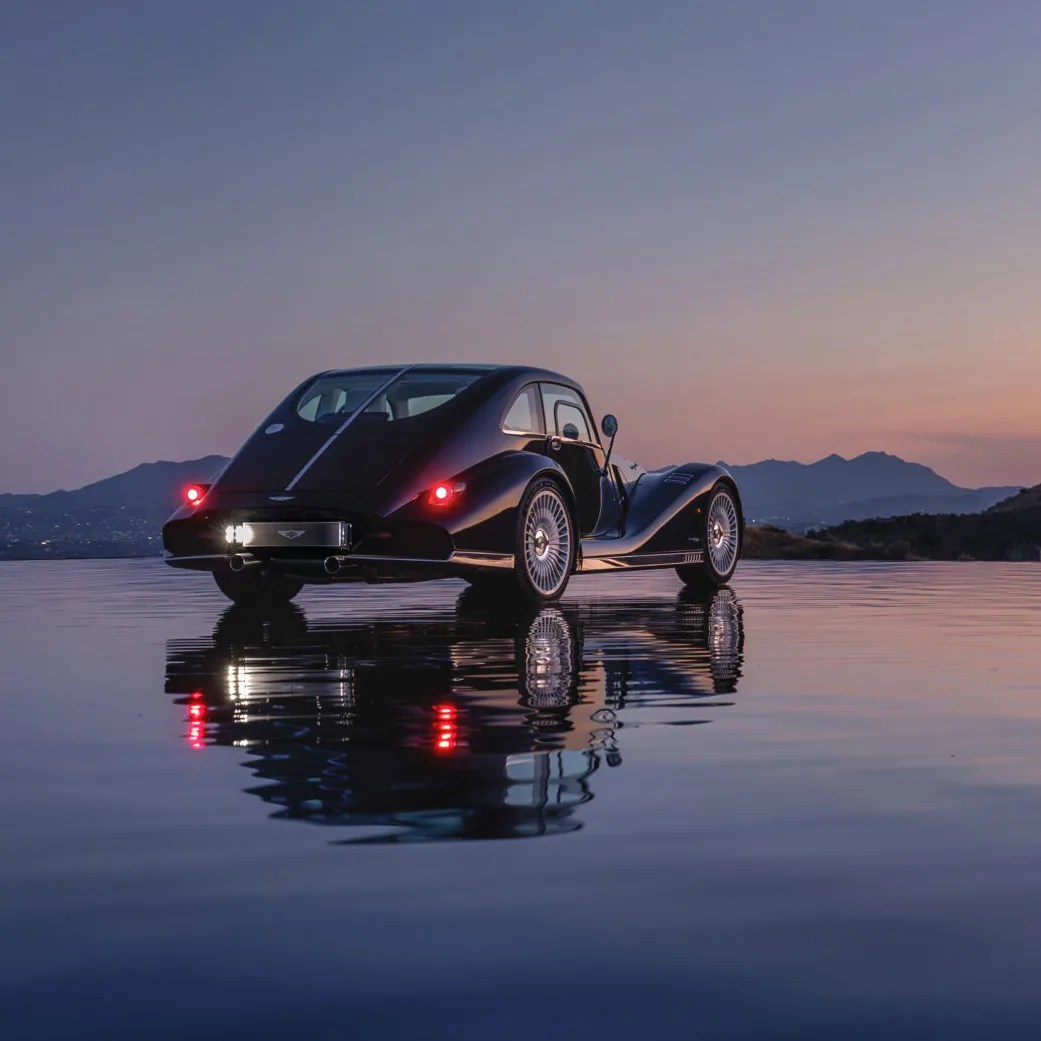 Black vintage-style car with round rear lights reflected on a calm water surface at dusk.