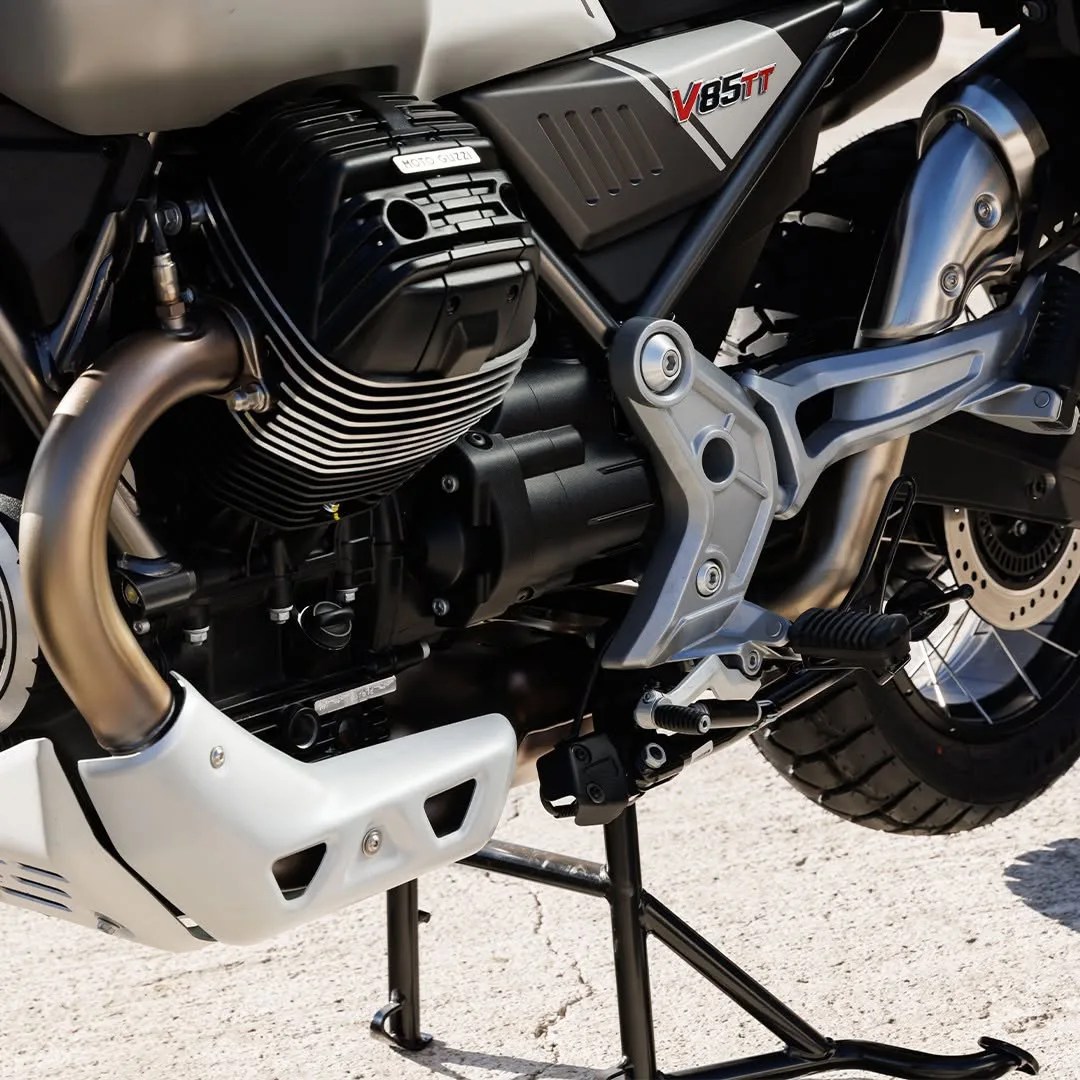 Close-up of a Moto Guzzi V85 TT motorcycle engine and exhaust system with black and silver components on a center stand.