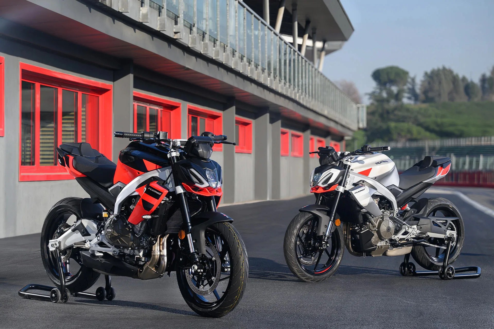 Two Aprilia Tuono motorcycles, one black and red and the other white and red, parked on a racetrack with red-framed windows in the background.