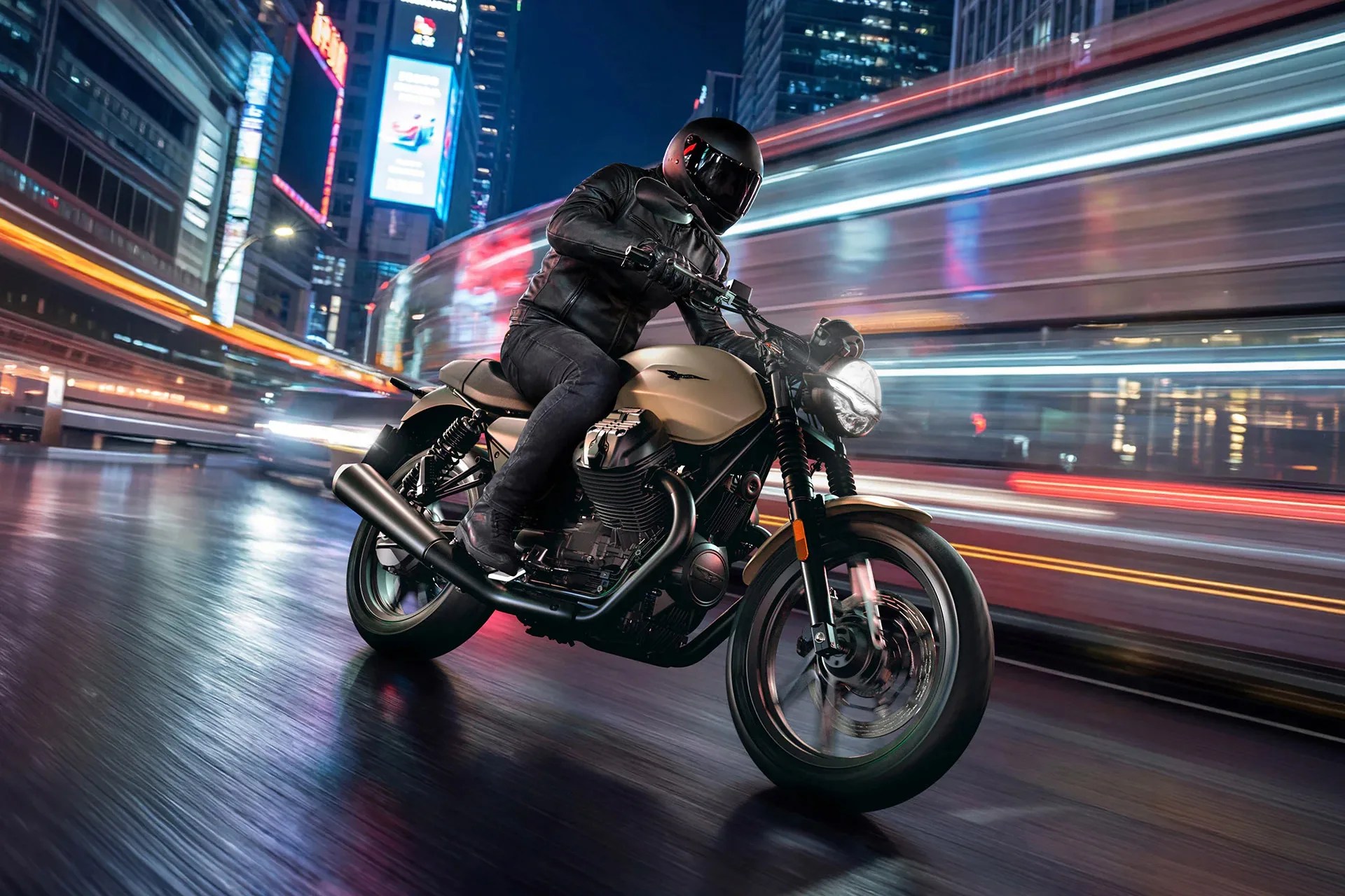 Motorcyclist in black gear riding a beige motorcycle on a wet city street at night with light trails.